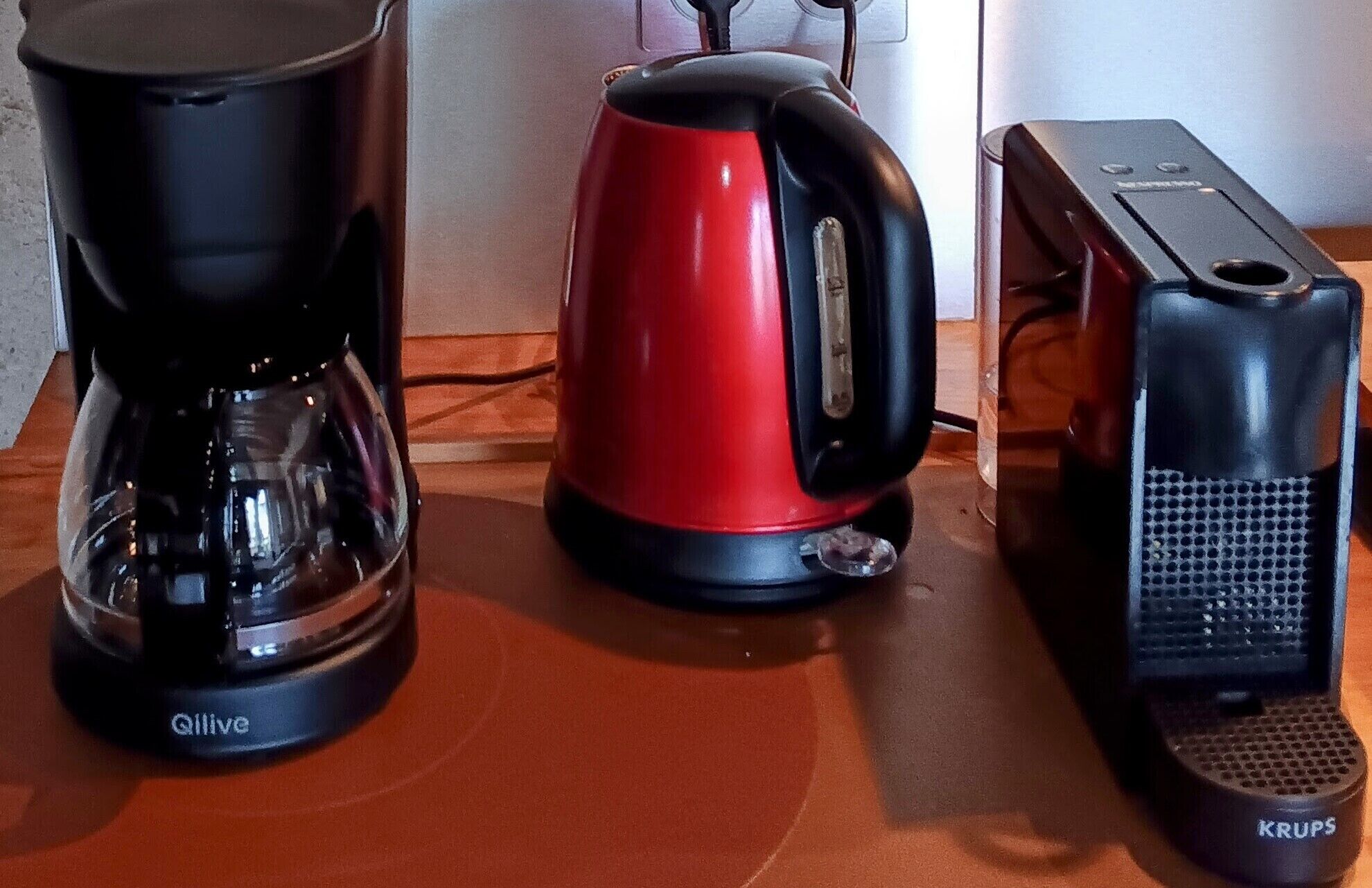 Kahvinkeitin, punainen sähköinen vedenkeitin ja Krups-merkkinen kahvimylly keittiötasolla.