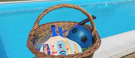 A wicker basket with a colorful ball and a blue paddle by a swimming pool.