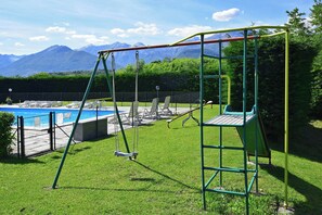 A playground with swings and a slide, a pool, and mountains in the background.