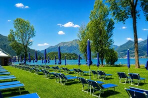 A row of blue lounge chairs and umbrellas on a grassy area with a lake and mountains in the background.