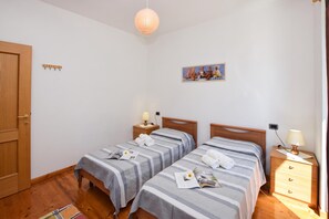 Habitación de hotel con dos camas individuales, una puerta de madera, una mesita de noche pequeña con lámpara, un cuadro colgado en la pared y suelo de madera.