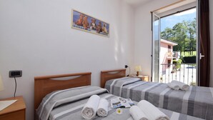 Una camera d'albergo con due letti, una finestra che dà su un balcone e alberi, e un quadro appeso al muro.