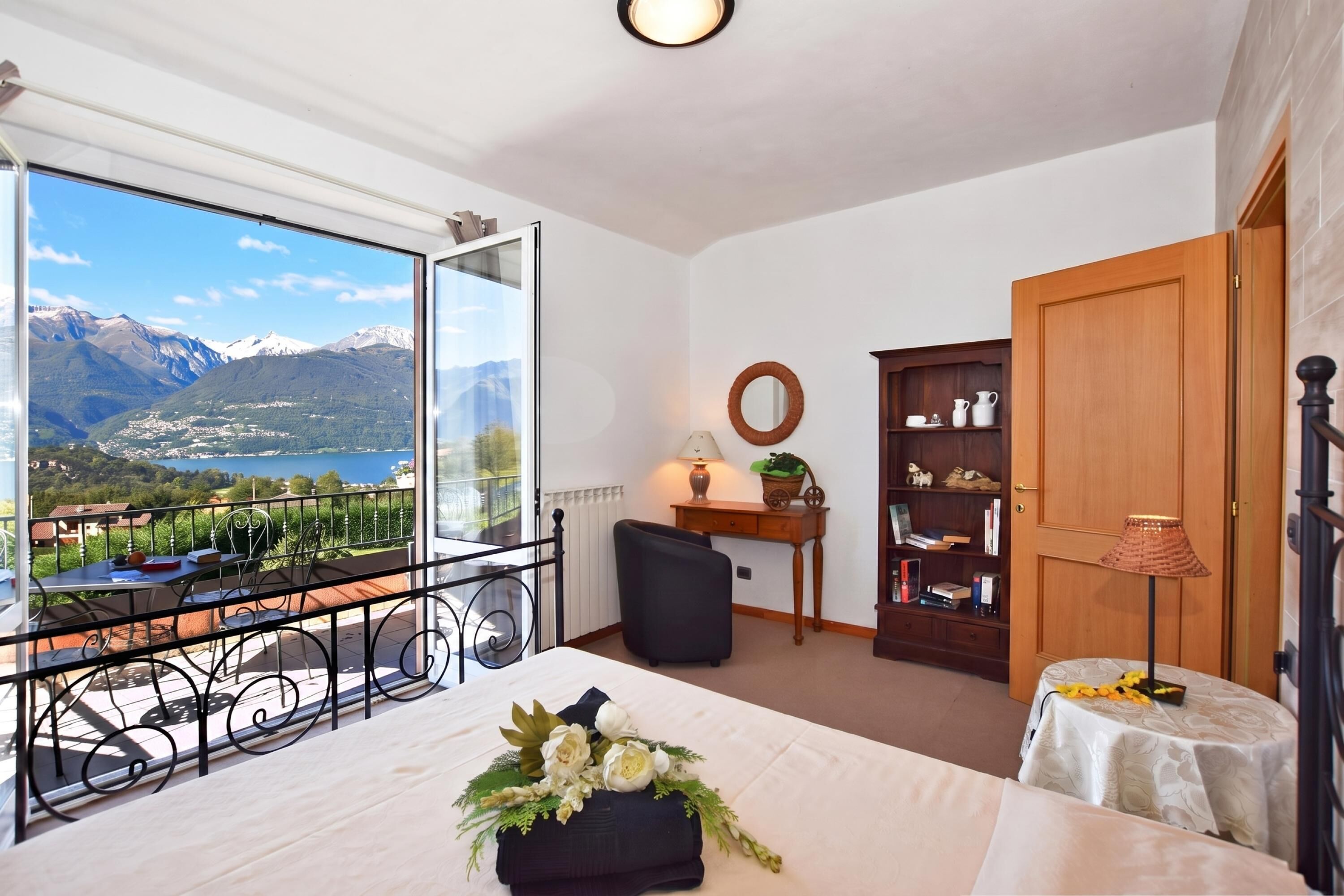Una camera da letto con un balcone, un letto, un tavolino con una lampada, una libreria e vista sulle montagne e su un lago.