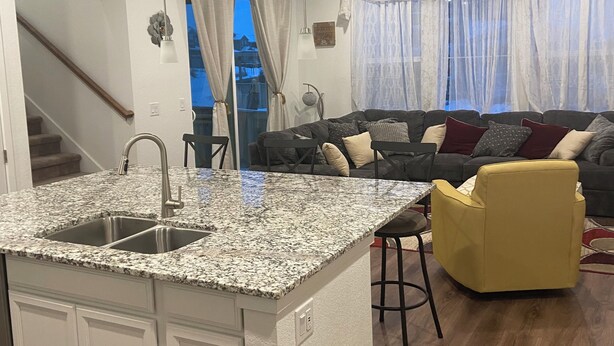 A modern kitchen with granite countertops, a built-in sink, and a small island. There is a yellow armchair and a dark-colored sofa in the living area. - Beautiful 2022 Constructed Patio Home. Sleeps up to 6 people. 10mins DIA Airport (Commerce City)