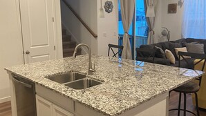 A kitchen with granite countertops, stainless steel sink, and white cabinetry. There is a dining area with a bar stool and a couch. - Beautiful 2022 Constructed Patio Home. Sleeps up to 6 people. 10mins DIA Airport (Commerce City)