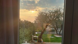A view through a glass door of a garden with a small table and chairs, a tree, and a statue. - House of Olives (Lazise)