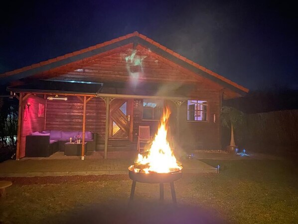 A wooden cabin with a fire pit in the foreground, a covered patio area with seating, and a visible window.