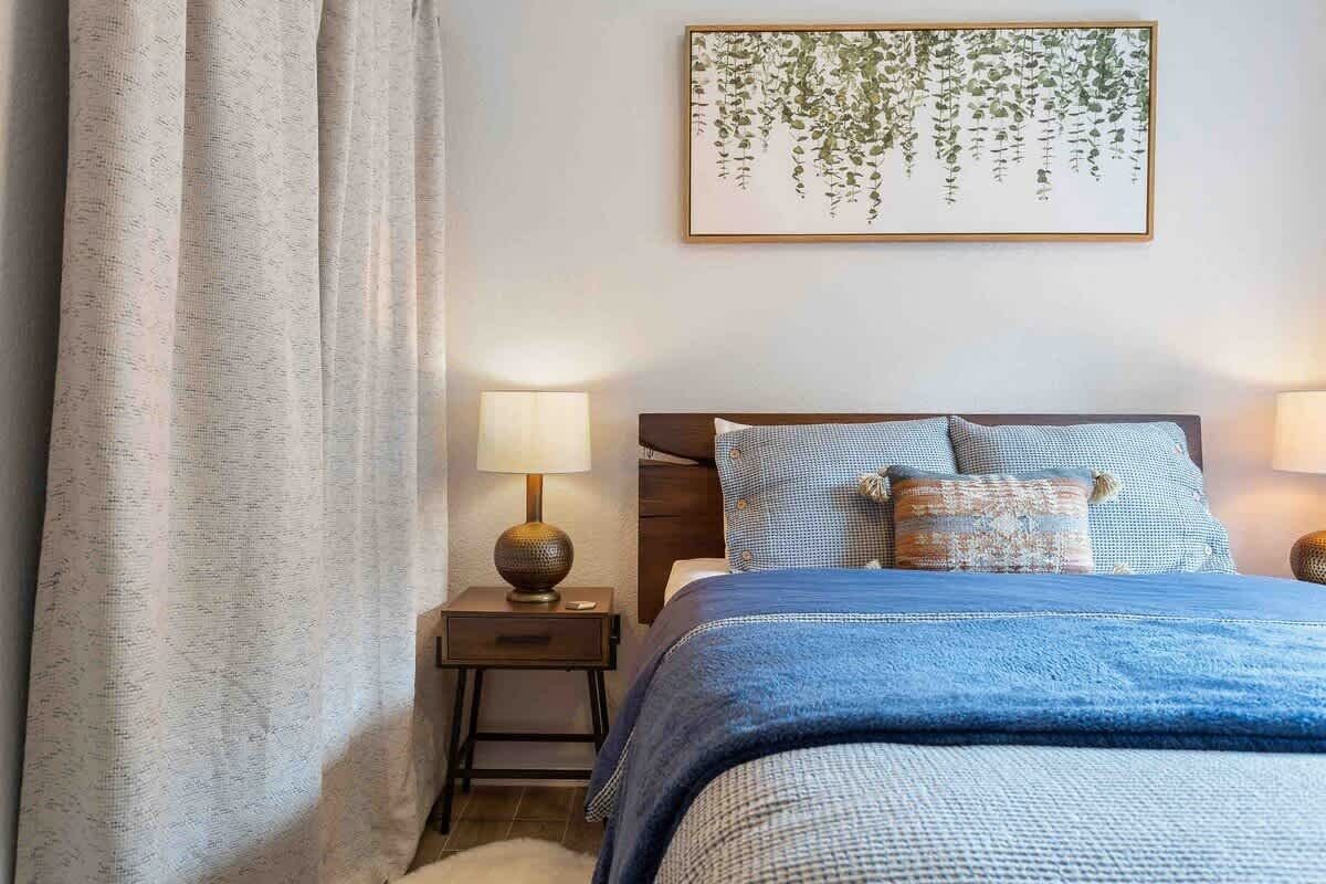 Une chambre à coucher avec un lit, deux lampes de chevet, une table de chevet, une plante en pot et un rideau.