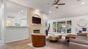 A spacious living room with a fireplace, a ceiling fan, and a large window providing a view of the outdoors. - 5BR/CedarPark/Soothing Sauna/HotTub/Peloton (Cedar Park)