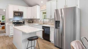 A modern kitchen with white cabinets, stainless steel appliances, and a central island with stools. - 5BR/CedarPark/Soothing Sauna/HotTub/Peloton (Cedar Park)