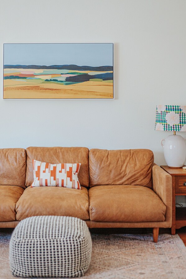 Un canapé en cuir marron agrémenté d’un coussin à motifs, un pouf à carreaux, un vase blanc avec un couvercle à carreaux et, au mur, un tableau représentant un paysage encadré.