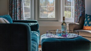 Een woonkamer met een bank, een poef en een glazen tafel met een fles en een glas.