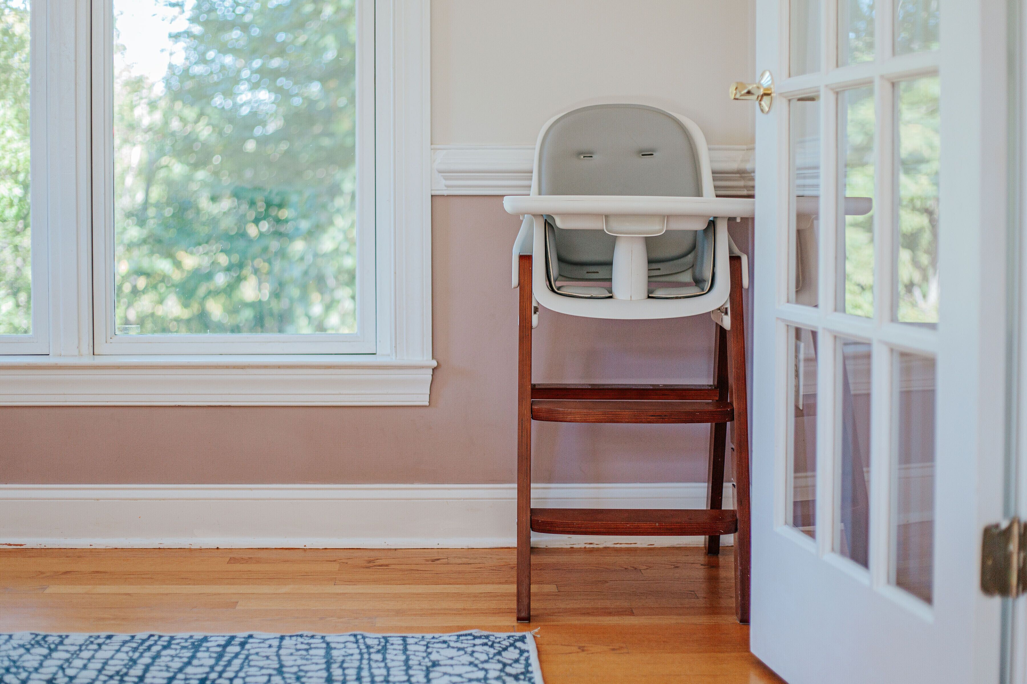 A room with a wooden floor, a window with a view of trees, a white door, and a white chair with a tray.