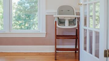 A room with a wooden floor, a window with a view of trees, a white door, and a white chair with a tray.