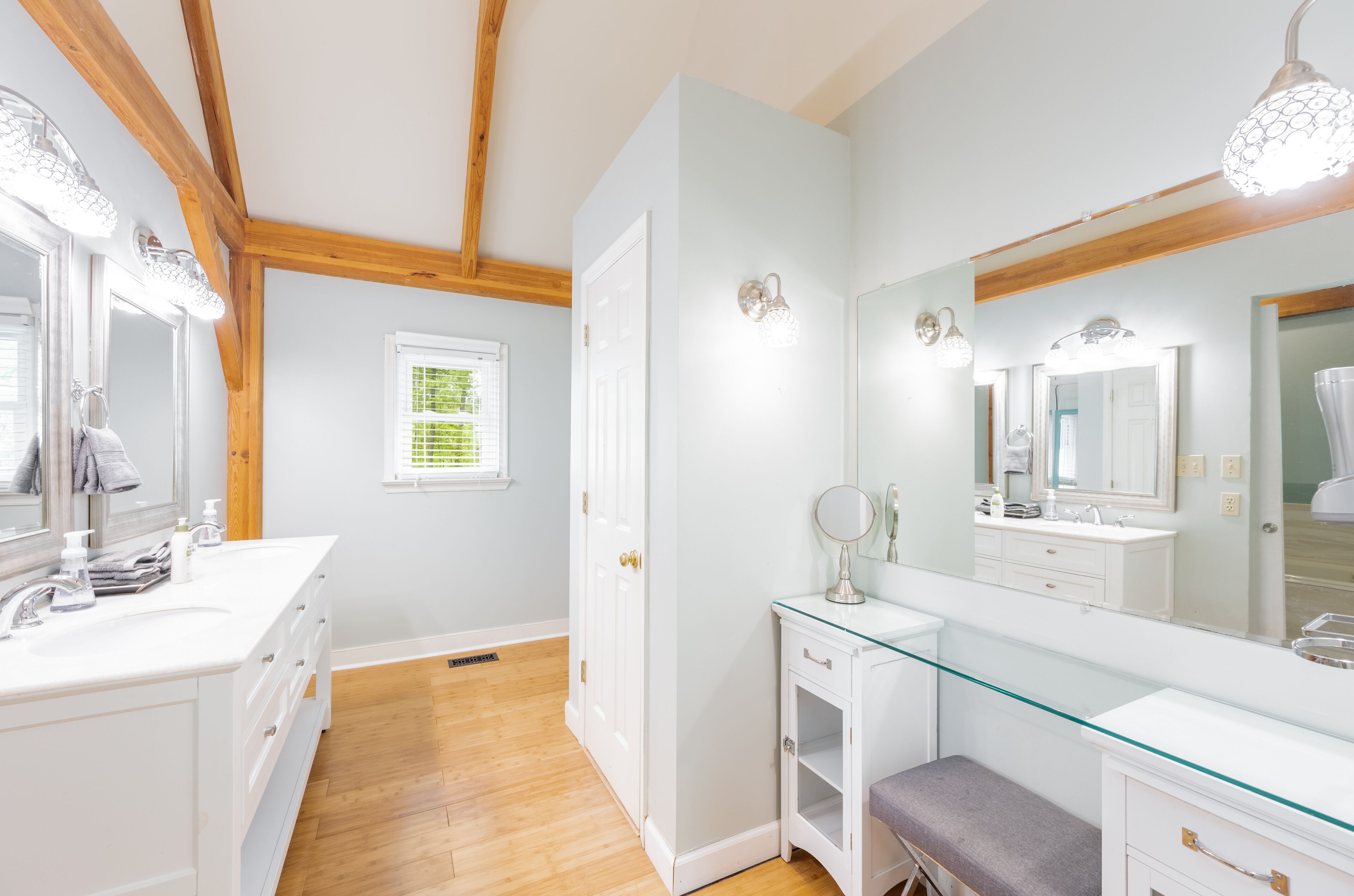 A bathroom with a white vanity, a glass-topped sink, and a large mirror. There is a door leading to another room and a window with blinds.