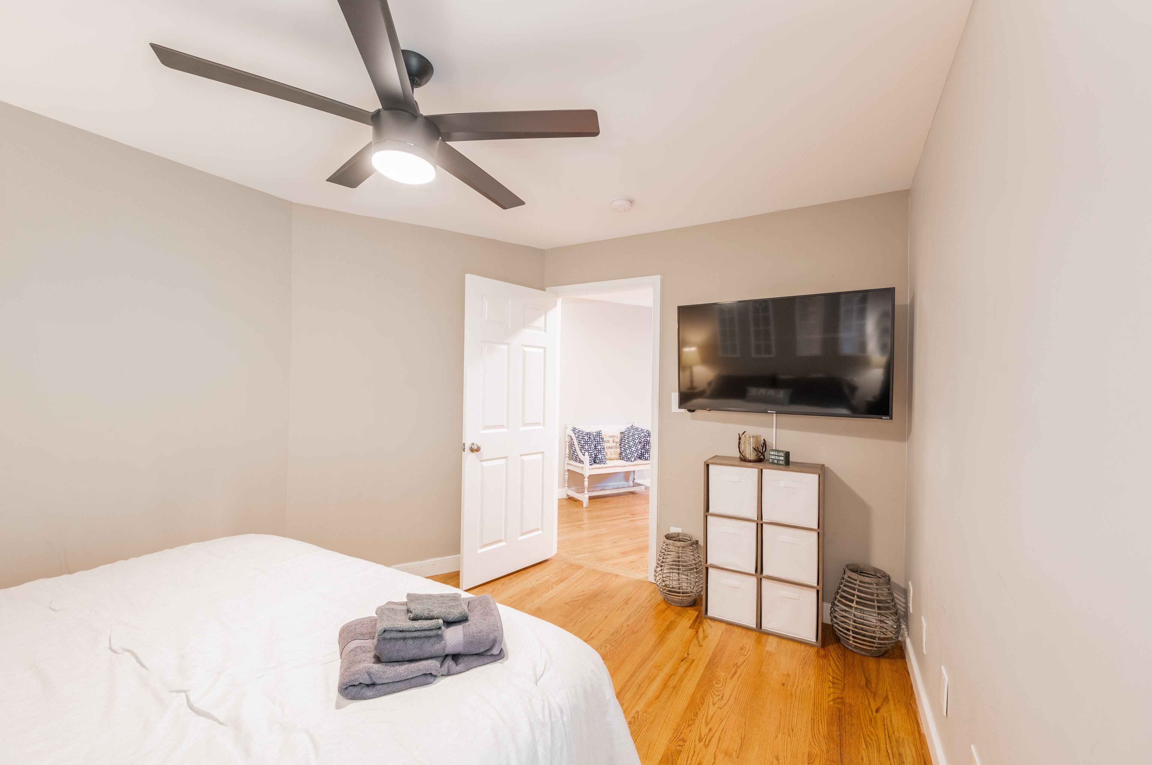 A bedroom with a bed, a ceiling fan, a TV mounted on the wall, and a door leading to another room.