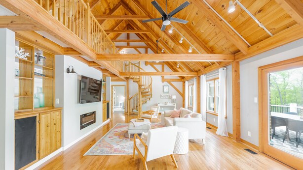 A spacious living area with wooden beams, a ceiling fan, a fireplace, and a large window overlooking a deck. - Beautiful and Private 6000 sqft waterfront home with private dock and hot tub! (Mineral)