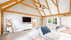 A spacious bedroom with a large bed, wooden ceiling beams, a ceiling fan, a TV, a dresser, and a window with a view of greenery. - Beautiful and Private 6000 sqft waterfront home with private dock and hot tub! (Mineral)