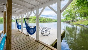 A wooden deck with a hammock and a chair overlooking a calm lake, surrounded by trees. - Beautiful and Private 6000 sqft waterfront home with private dock and hot tub! (Mineral)