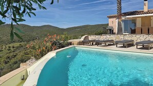 A pool with lounge chairs, a stone wall, and a view of green hills.