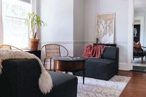 Un salon avec un canapé foncé, une table basse ronde, un fauteuil en osier, une plante en pot et un grand tableau abstrait accroché au mur.