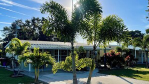 Un jardín bien cuidado con palmeras y plantas con flores, un camino empedrado y un edificio con tejado de tejas al fondo.