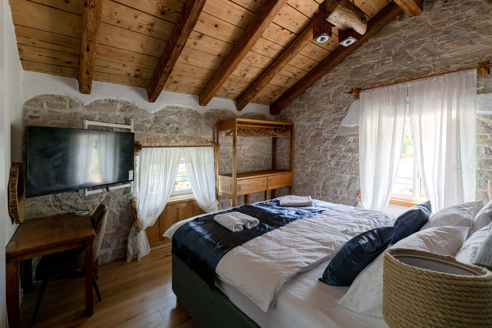Une chambre rustique avec un mur en pierre, des poutres apparentes en bois au plafond, un lit avec des oreillers, une table de chevet en bois, une télévision et une fenêtre ornée de rideaux légers.