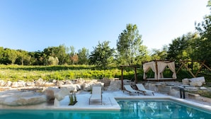 Ein Schwimmbecken mit Liegestühlen, eine Holzpergola mit Dach und eine Steinmauer.