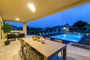 Área de piscina externa com espreguiçadeiras, mesa de jantar e vista do bairro ao entardecer.