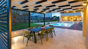 En moderne udendørs terrasse med swimmingpool, et spiseområde med stole og udsigt til en boligbygning.