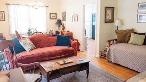A cozy living room with a red sofa, a wooden coffee table, and a beige armchair. There is a chandelier hanging from the ceiling, and a door leading to another room. - Newly renovated, comfy & artsy! 1mi to Square, 2mi to Campus in old Oxford (Oxford)