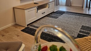 Ein Wohnzimmer mit Fernseher auf einem Holzschrank, einer kleinen Pflanze auf einem Regal und einem Essbereich mit Tisch und Stühlen.
