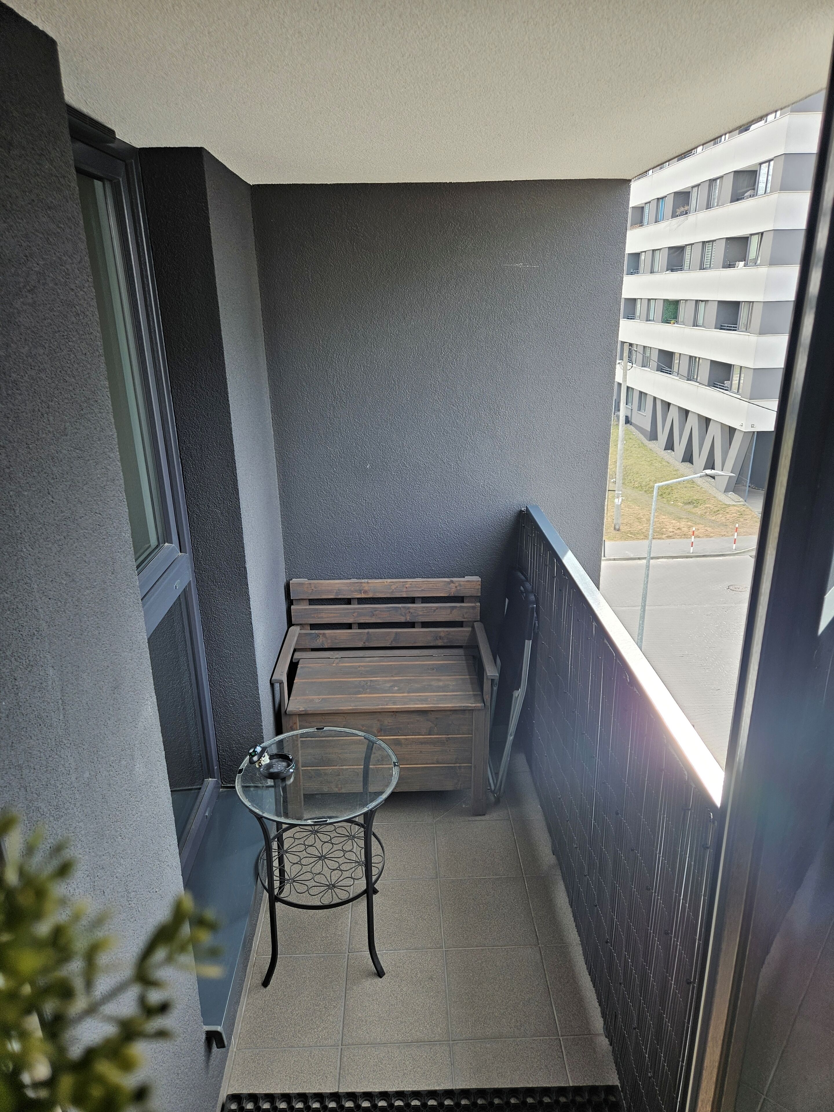 A balcony with a wooden bench, a glass table, and a view of a building across the street.