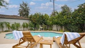 A poolside area with wooden lounge chairs and a table, surrounded by trees and a fence. - ❤️of PHX in Historic Willo • Heated Pool • Chef's Kitchen • Fast WiFi (Phoenix)