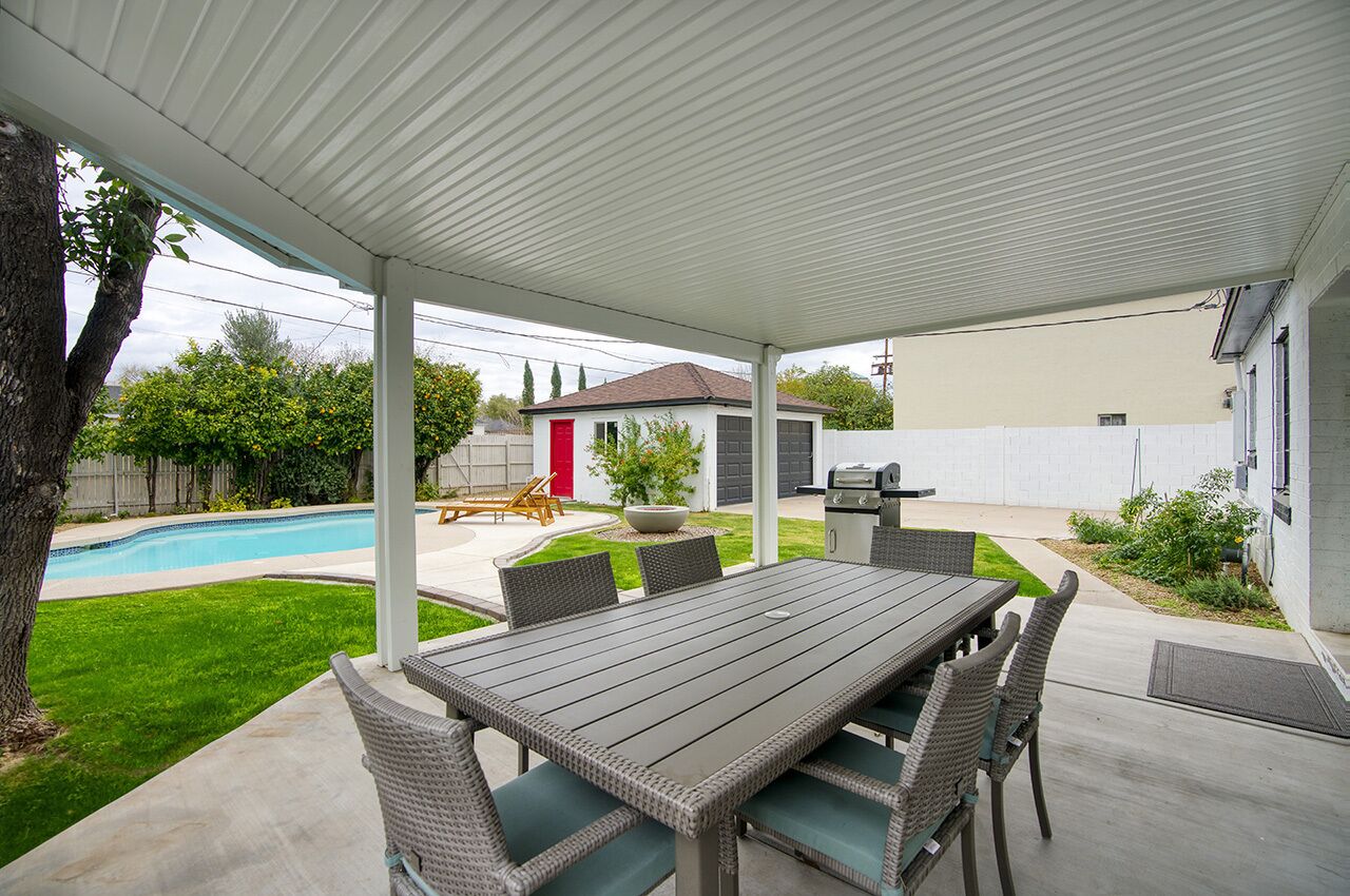 Un porxo cobert amb taula i cadires, una barbacoa i una piscina al fons.