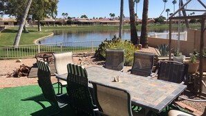 An outdoor patio with a table and chairs, a fire pit, and a view of a lake and palm trees. - Lakefront Home fully furnished (Sun Lakes)