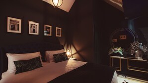 A dark-themed bedroom with a bed, bedside lamps, framed pictures on the wall, and a vanity with a mirror and vase. - The Rockefeller Apartments (Windermere)