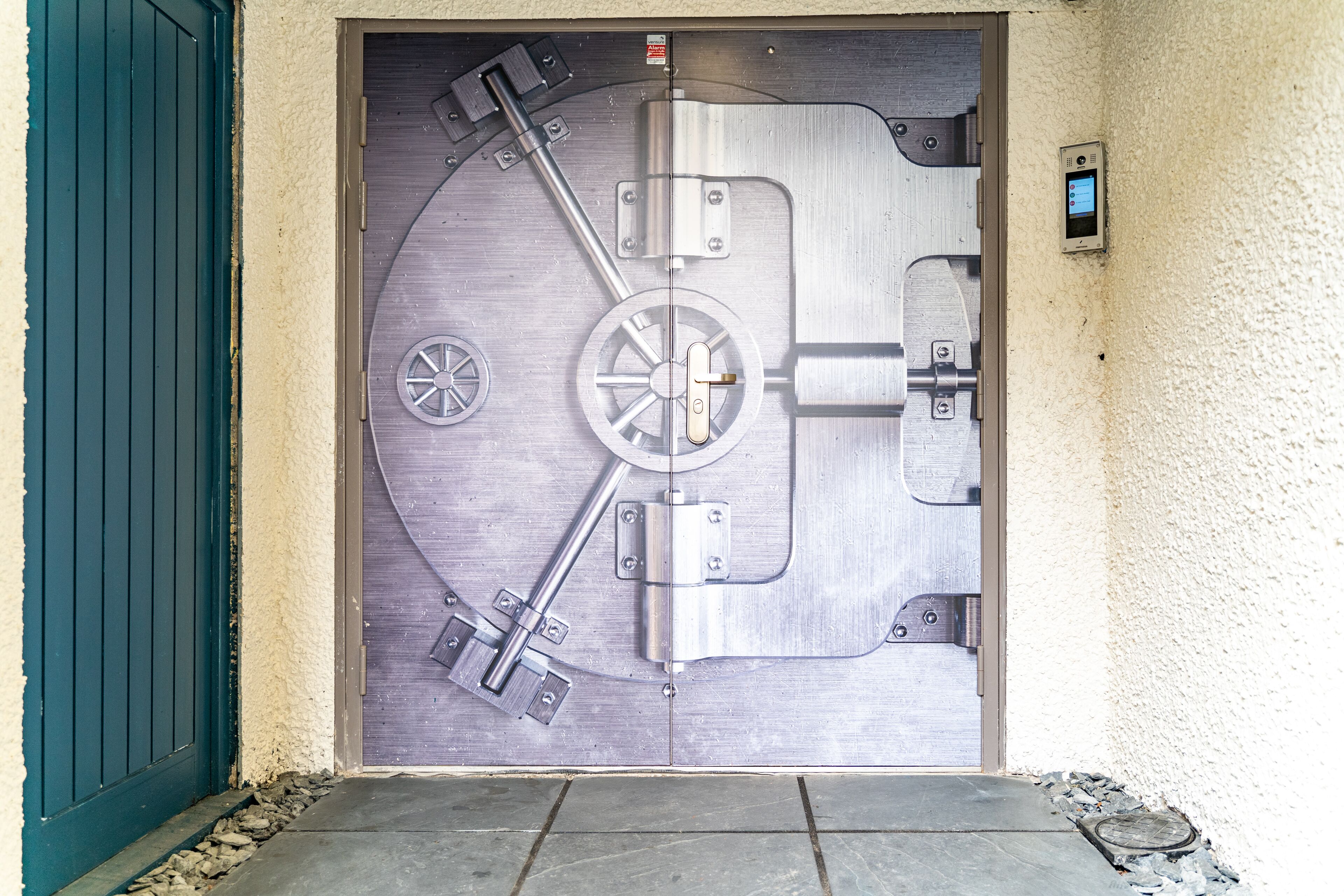 A large, metallic vault door with a complex locking mechanism, a small security panel on the wall, and a green door frame.