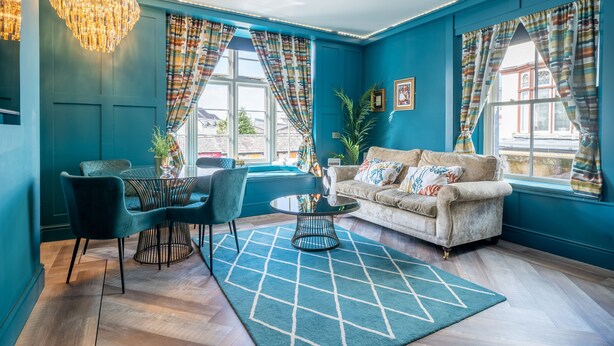 A room with teal walls, a beige sofa, a round coffee table, and a chandelier. - The Rockefeller Apartments (Windermere)