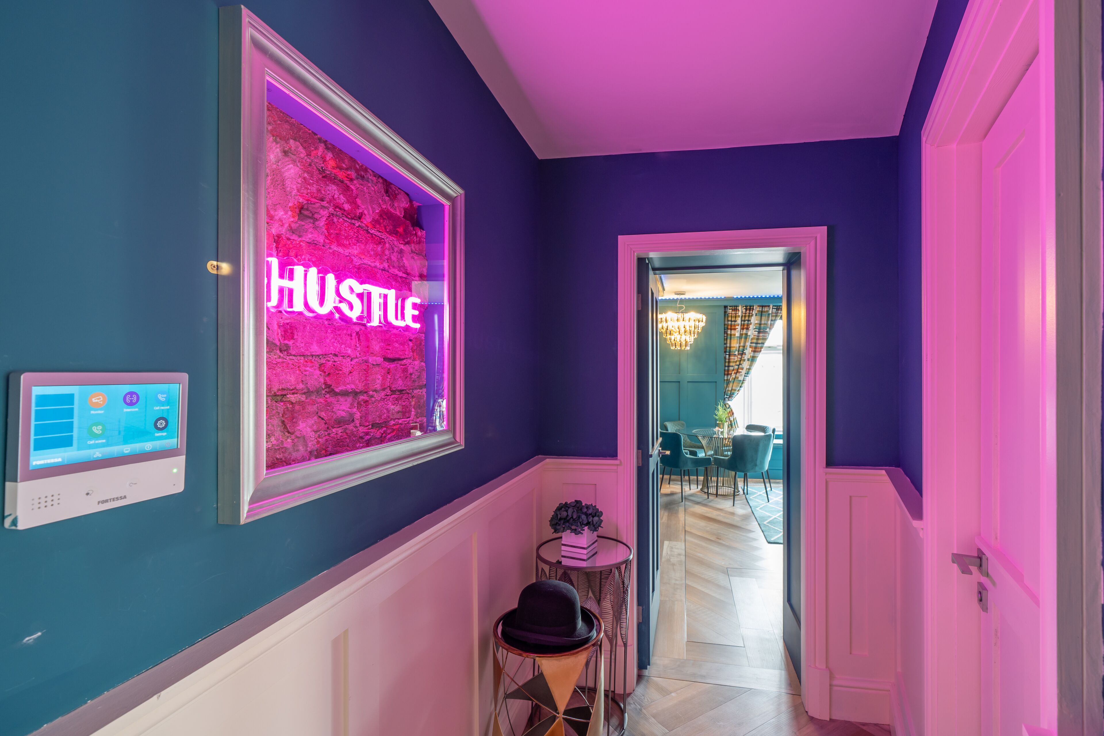 A hallway with a neon 'HUSTLE' sign, a thermostat, and a small table with a plant.