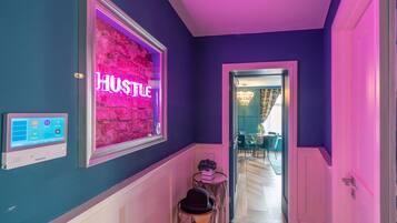 Un couloir avec un panneau néon indiquant le mot « HUSTLE », un thermostat et une petite table sur laquelle est posée une plante.