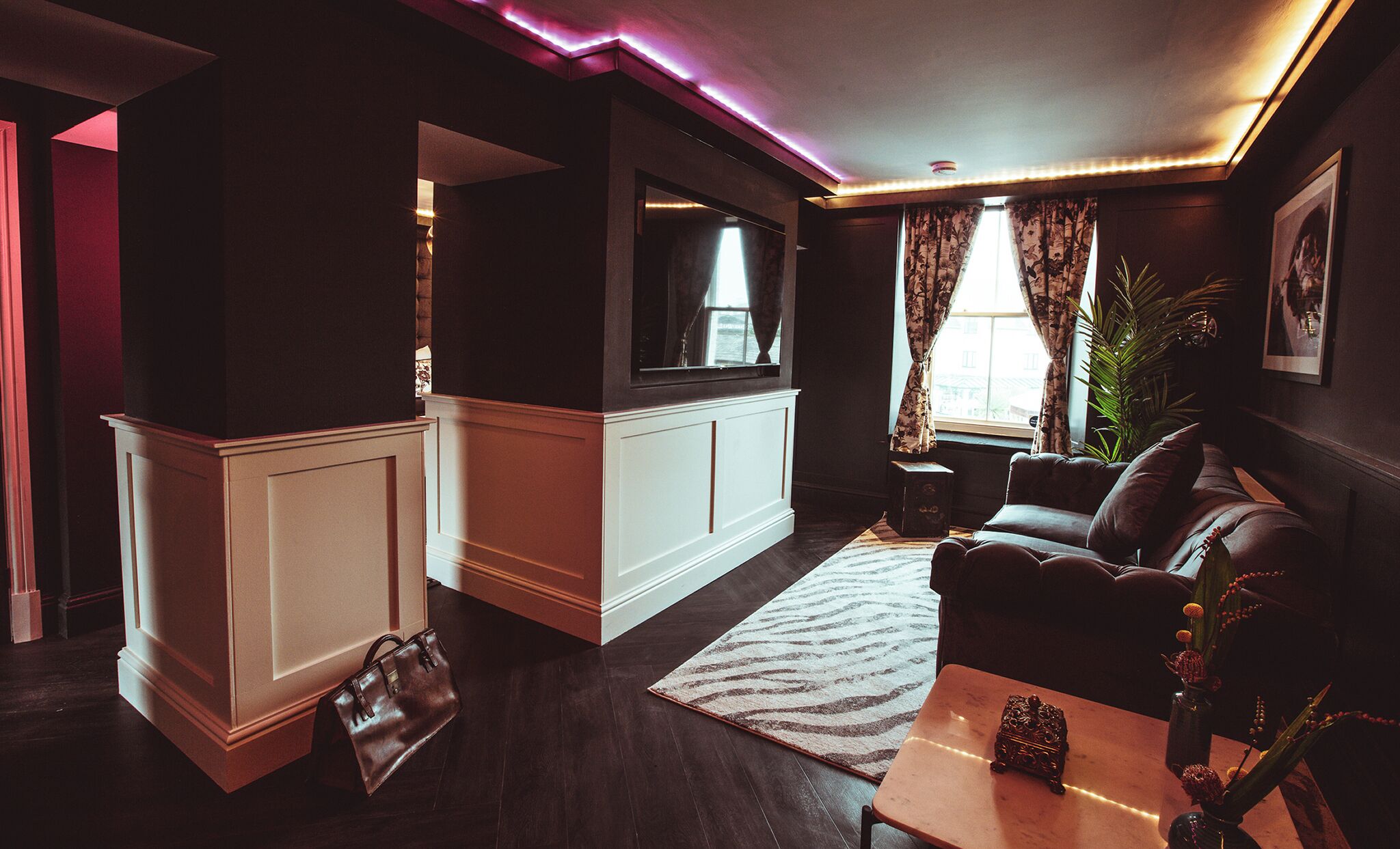 A dimly lit living room with dark wood flooring, a leather sofa, a small coffee table, and a TV mounted on the wall.