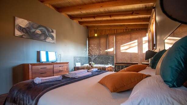 A hotel room with a bed, wooden ceiling, artwork on the wall, and a desk with a computer.