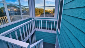 Un coin d’un bâtiment avec un revêtement de façade de couleur turquoise et des balustrades blanches, agrémenté d’une grande fenêtre offrant une vue sur une maison et des arbres.
