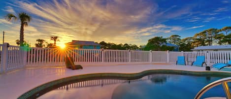 A swimming pool with a white fence, palm trees, and a sunset in the background.