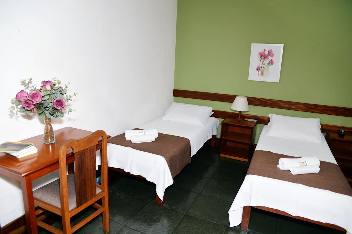 Quarto de hotel com duas camas de solteiro, uma escrivaninha de madeira com cadeira, um vaso com flores e um quadro na parede.