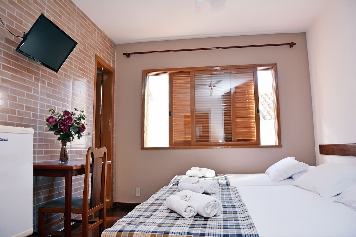 Quarto de hotel com cama, escrivaninha com cadeira, televisão fixada na parede e janela com persianas.