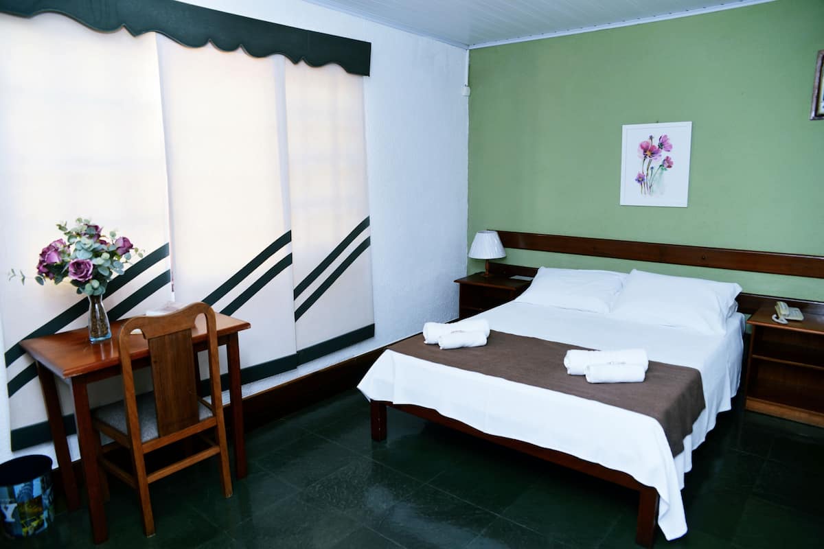 Quarto de hotel com cama, escrivaninha e cadeira, vaso com flores e um quadro na parede.