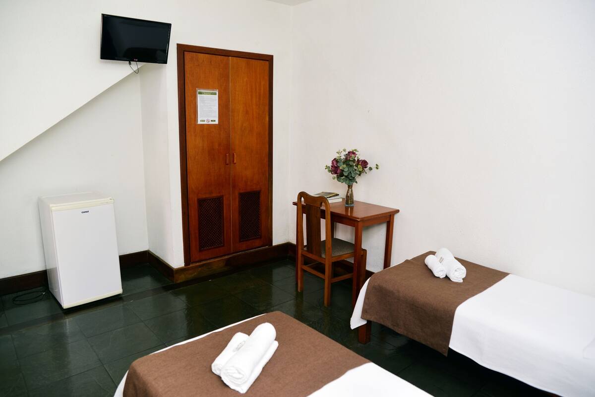 Quarto de hotel com duas camas, uma mesinha com um vaso, uma televisão na parede e um refrigerador.