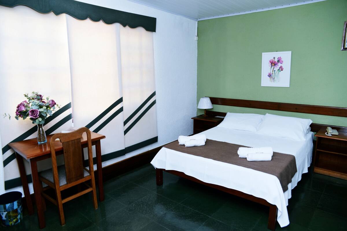 Quarto de hotel com cama, escrivaninha, cadeira, abajur e um vaso com flores.